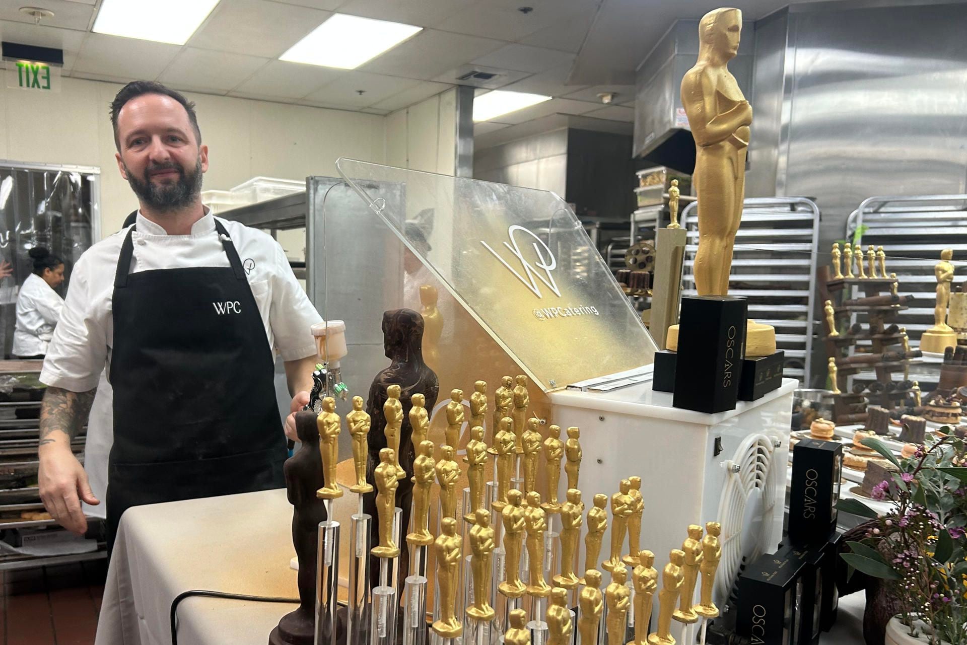 Un ayudante de cocina posa junto a figuras de chocolate de la estatuilla de los premios Oscar este jueves, en las cocinas del 